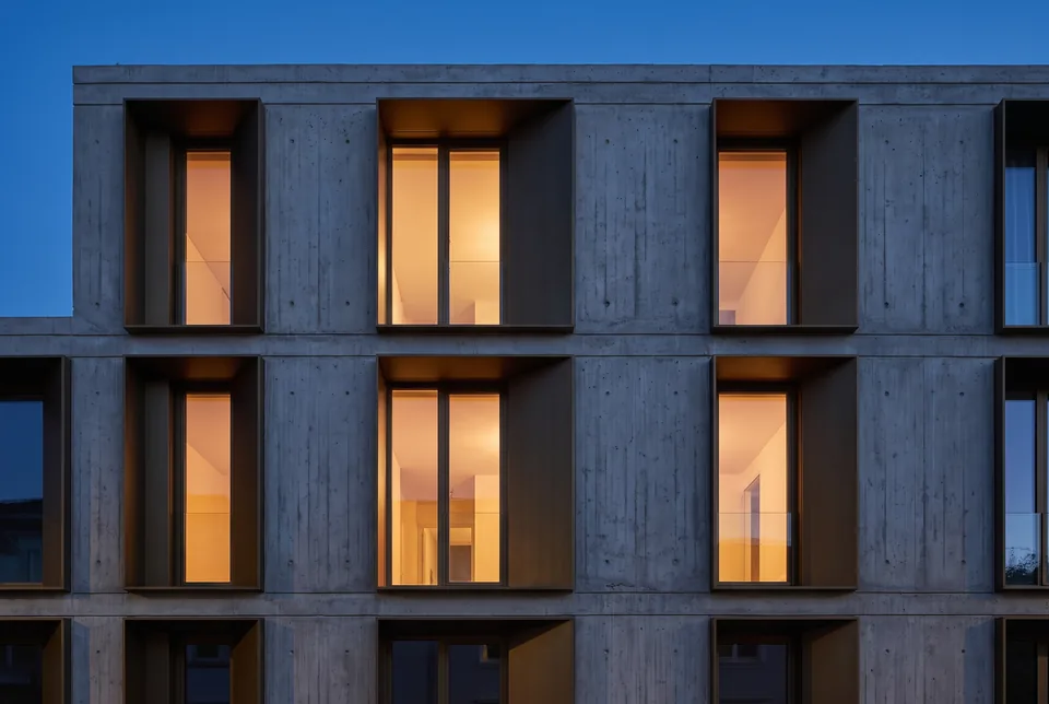 Board-formed concrete facade with bronze window reveals and lit interiors behind tall glazing at dusk