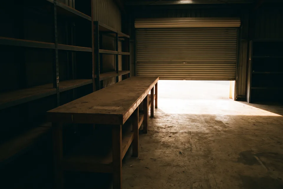 An empty workbench and steel shelving lit by a half-open roller shutter